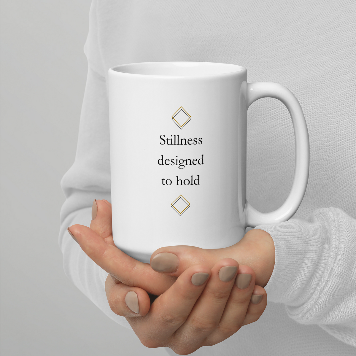 White ceramicmug with the quote "Stillness designed to hold" and subtle geometric details,held in both hands.